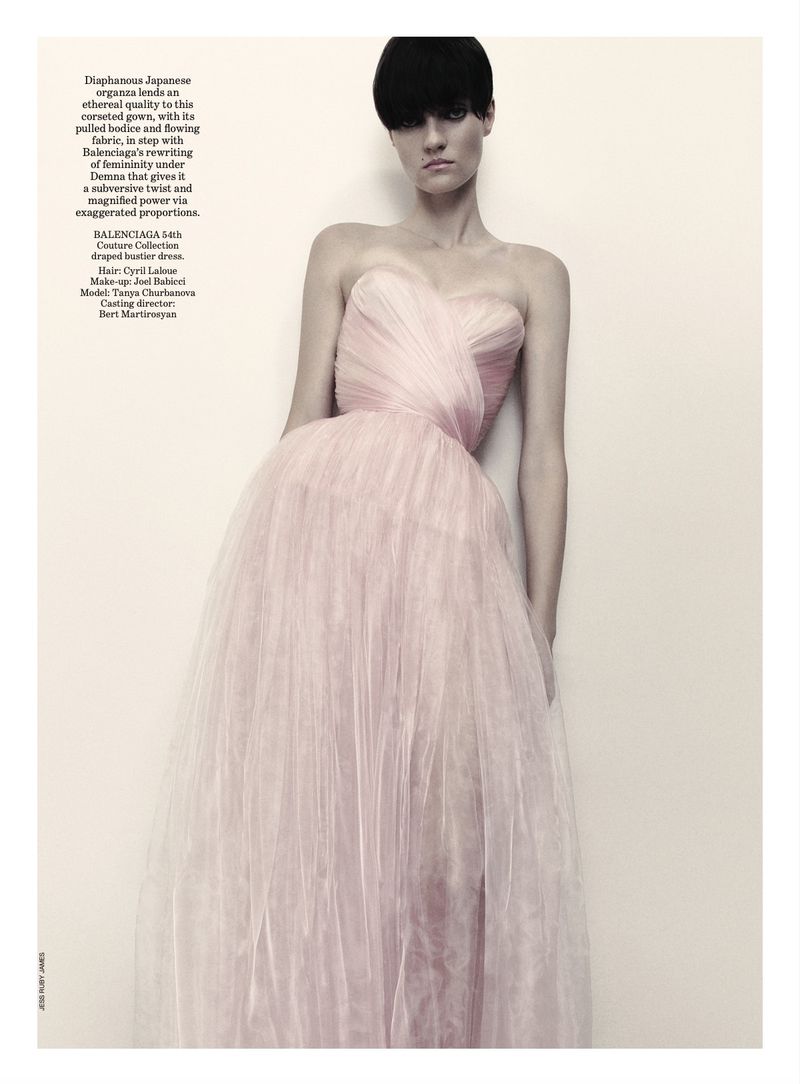 Editorial fashion shot of a woman with short black hair wearing a pink strapless tulle gown standing against a light neutral-colored wall.