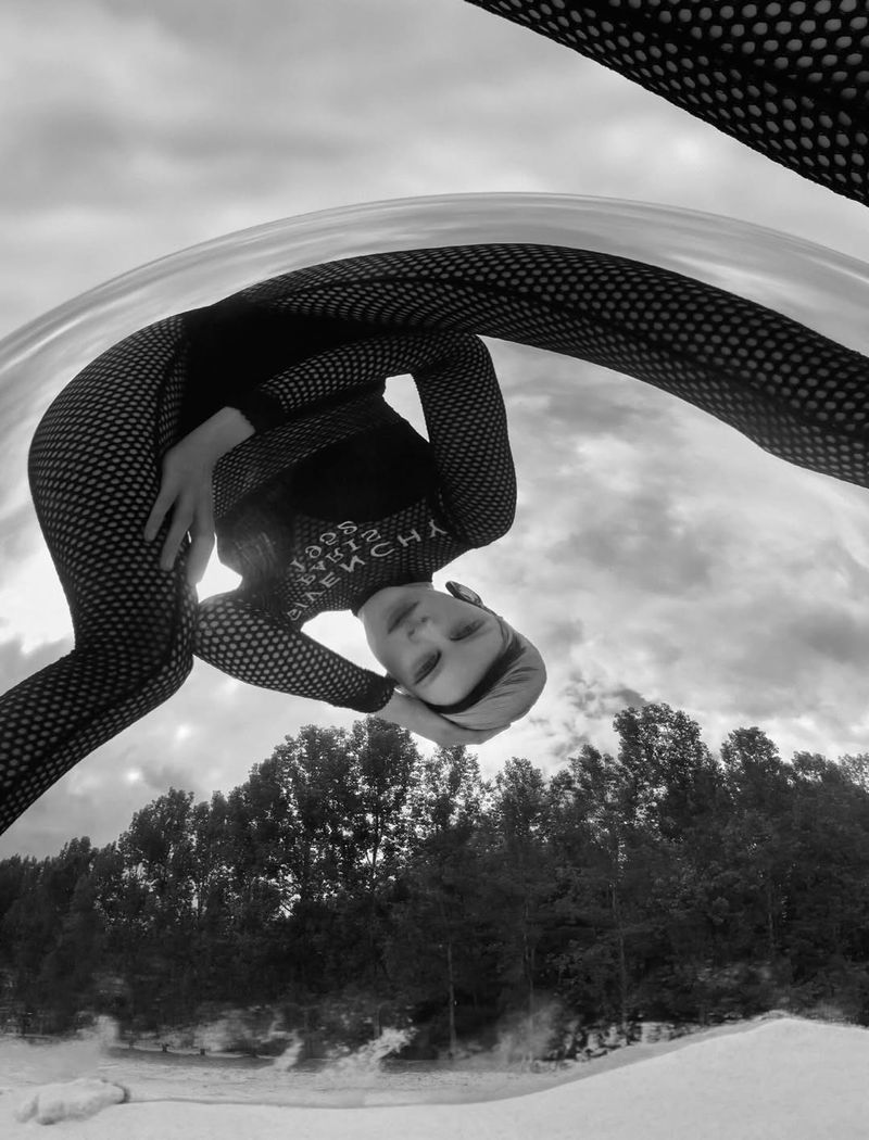 Monochrome portrait of an adult woman in a bodysuit, inverted perspective with a backdrop of trees and cloudy sky, artistic and surreal.