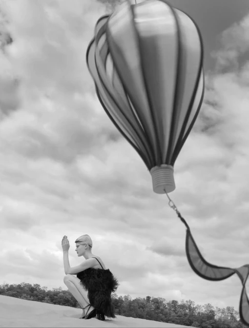 Artistic monochrome shot of a woman in a feathered dress crouching in a landscape, featuring a decorative hot air balloon.