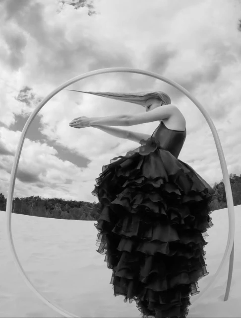 Elegant black and white portrait of a woman with long hair standing in a field with a round prop against a cloudy sky background.