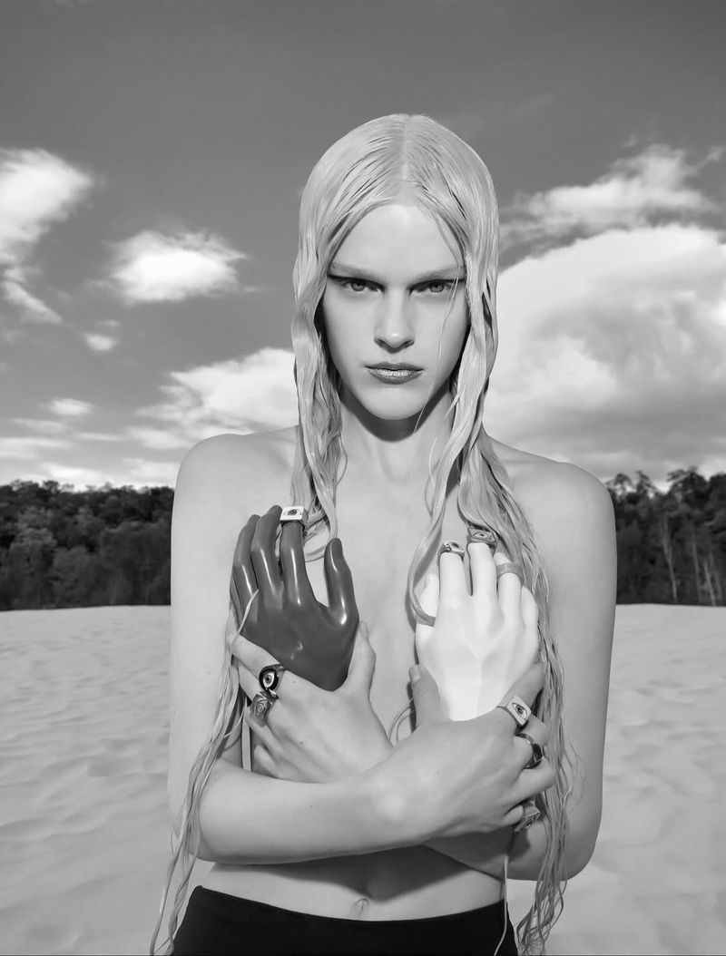 Black and white portrait of a striking woman with platinum blonde hair, dramatic makeup, adorned with jewelry, and set against a cloudy sky.
