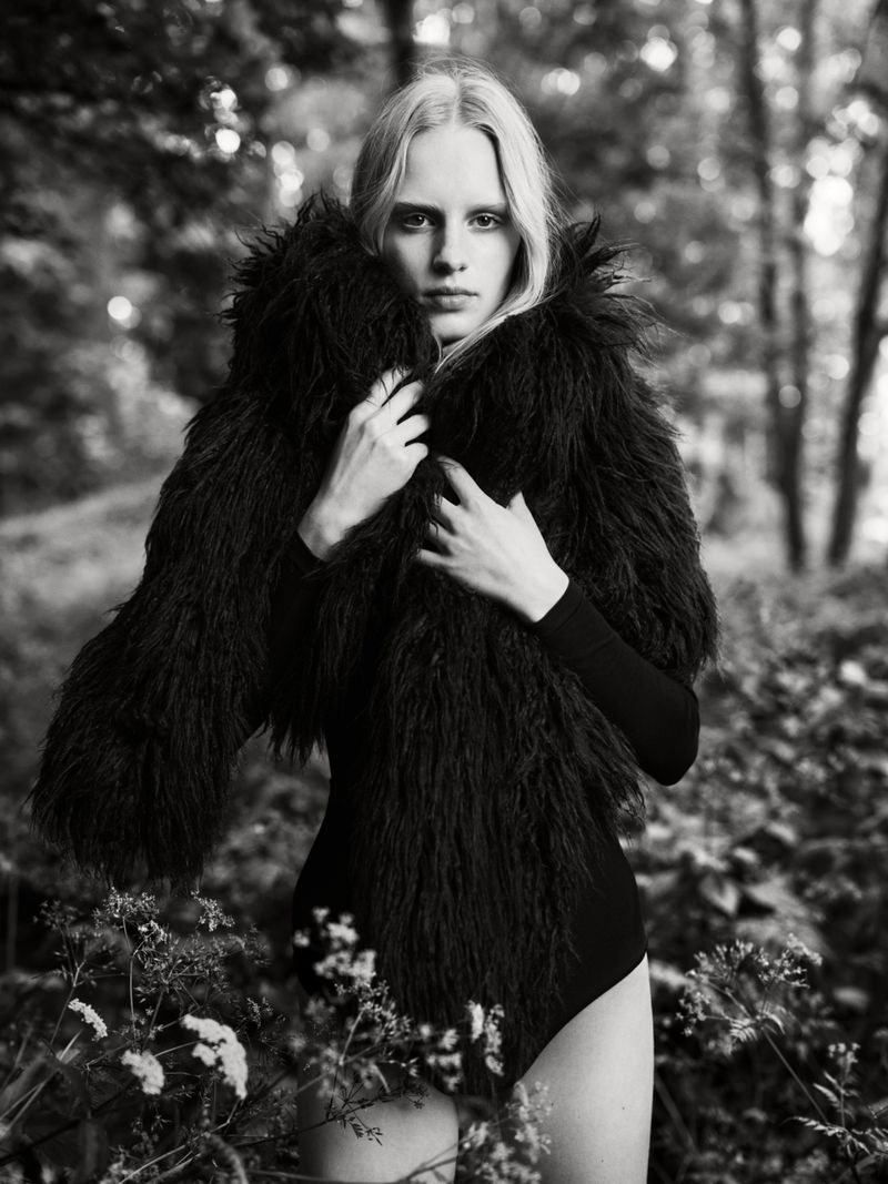 Monochrome portrait of a slender woman with long blonde hair, wearing a furry black jacket in an outdoor setting with blurred trees and flowers.