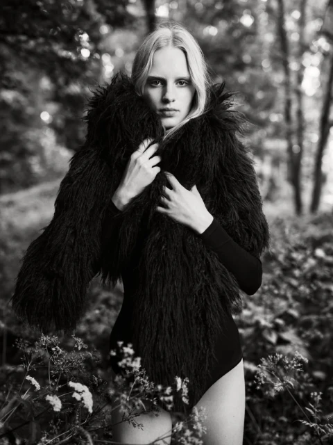 Monochrome portrait of a slender woman with long blonde hair, wearing a furry black jacket in an outdoor setting with blurred trees and flowers.