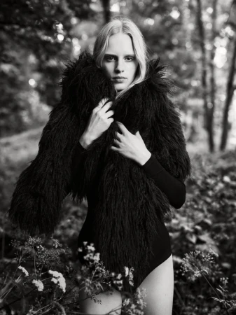 Monochrome portrait of a slender woman with long blonde hair, wearing a furry black jacket in an outdoor setting with blurred trees and flowers.