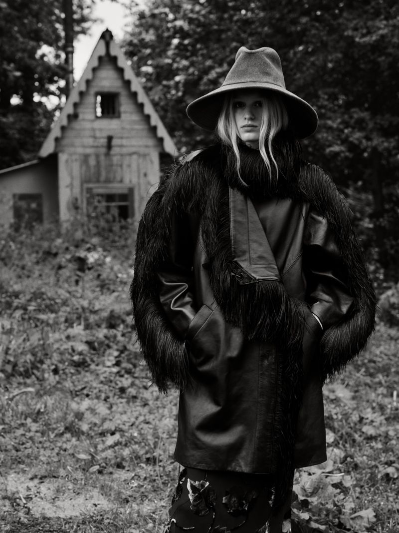 A woman with long blonde hair poses in a vintage hat and coat, standing outdoors against a backdrop of trees and a rustic wooden cabin.