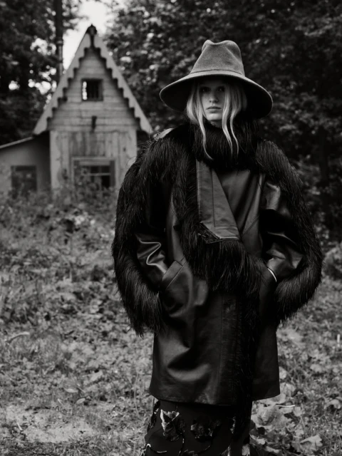 A woman with long blonde hair poses in a vintage hat and coat, standing outdoors against a backdrop of trees and a rustic wooden cabin.