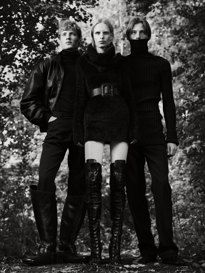 Three fashion models in black outfits pose in a monochromatic outdoor fashion shot with a forest background, showcasing elegant style.