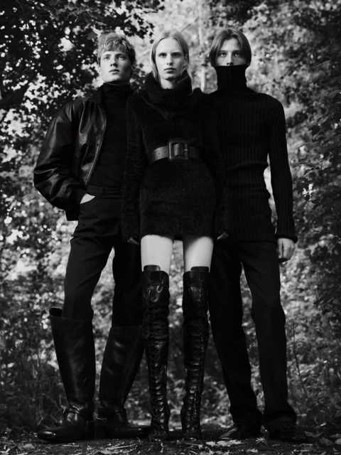 Three fashion models in black outfits pose in a monochromatic outdoor fashion shot with a forest background, showcasing elegant style.