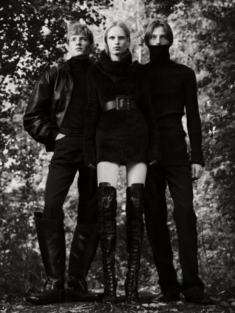 Three fashion models in black outfits pose in a monochromatic outdoor fashion shot with a forest background, showcasing elegant style.