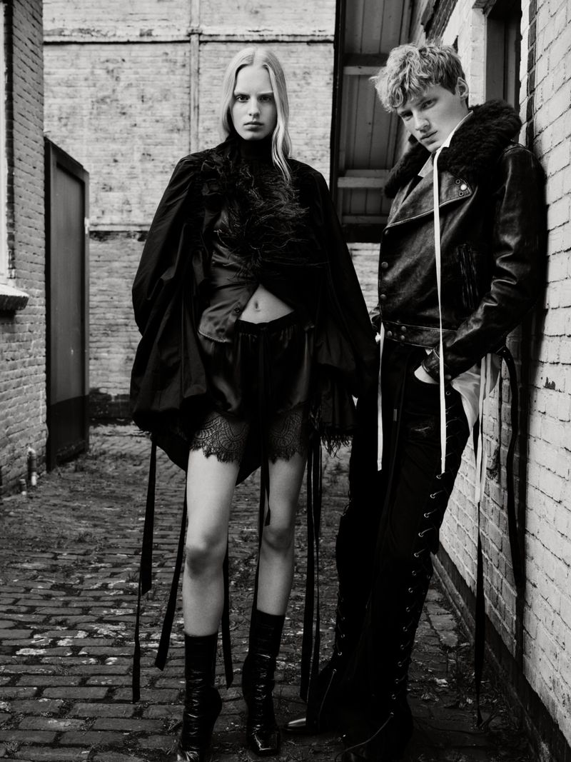 Black and white fashion photograph of a woman and man in dark clothing standing against brick walls in a narrow outdoor alley.