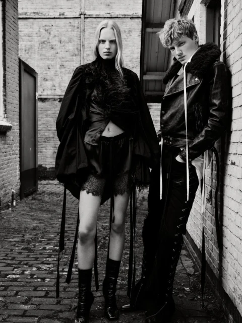 Black and white fashion photograph of a woman and man in dark clothing standing against brick walls in a narrow outdoor alley.