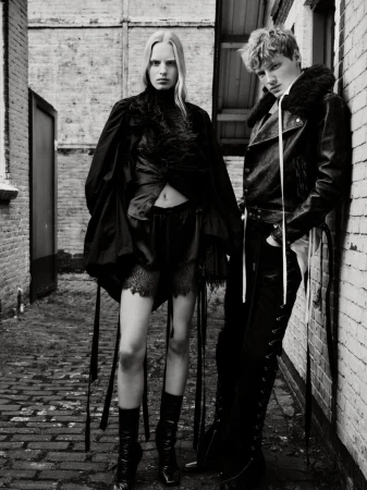 Black and white fashion photograph of a woman and man in dark clothing standing against brick walls in a narrow outdoor alley.