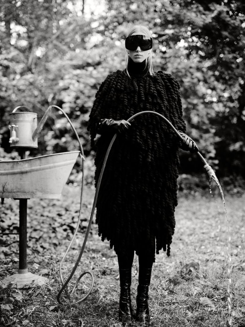 Black and white fashion shot of a woman in knitwear, dark glasses, and high boots, holding a garden hose outdoors with a watering can.