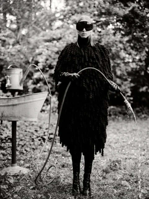 Black and white fashion shot of a woman in knitwear, dark glasses, and high boots, holding a garden hose outdoors with a watering can.
