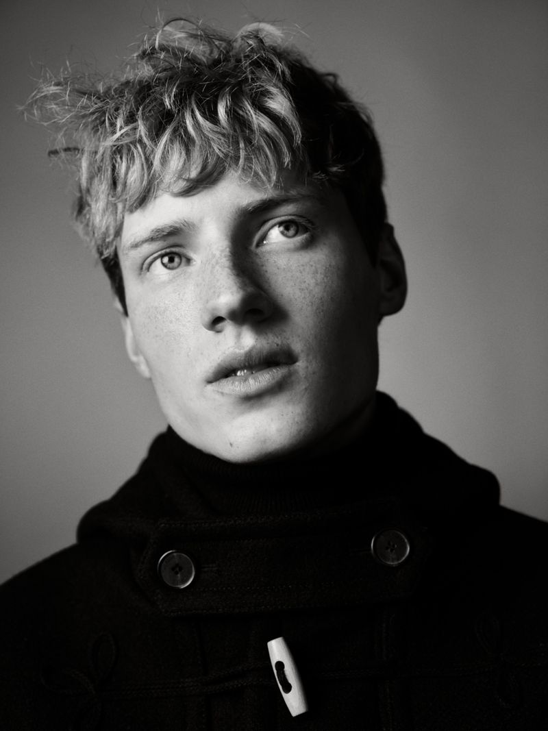 Black and white portrait of a young adult male with tousled blond hair and wearing a black duffle coat, shot in a photo studio.