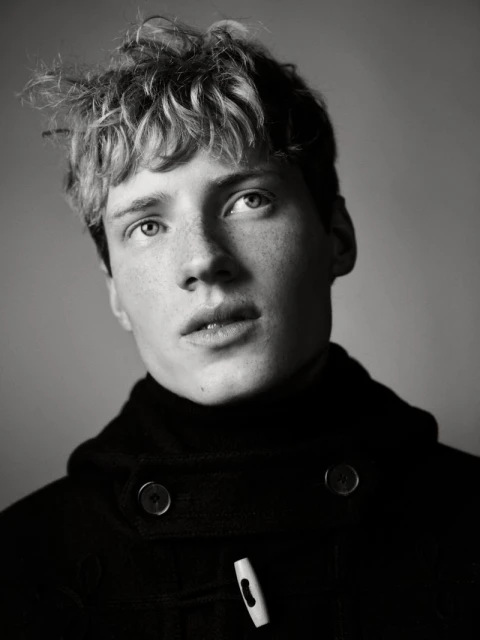 Black and white portrait of a young adult male with tousled blond hair and wearing a black duffle coat, shot in a photo studio.