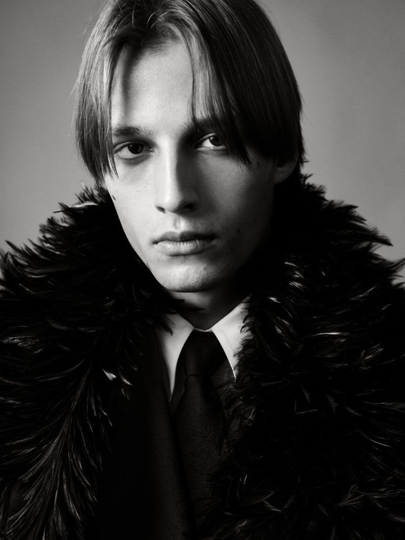 Monochrome portrait of a young adult male wearing a feather boa, collared shirt, and tie, set against a neutral background.