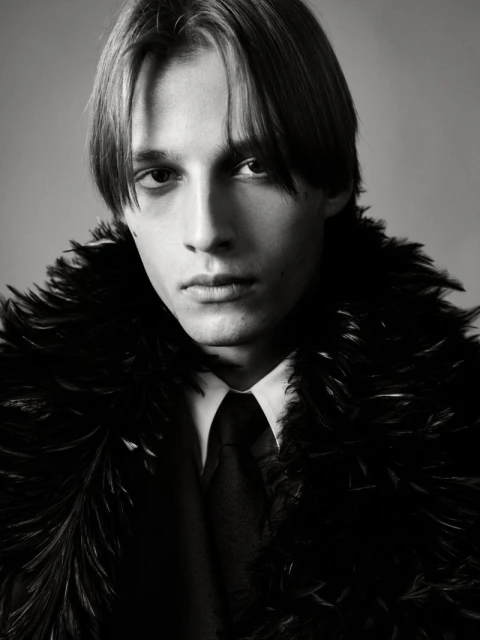 Monochrome portrait of a young adult male wearing a feather boa, collared shirt, and tie, set against a neutral background.