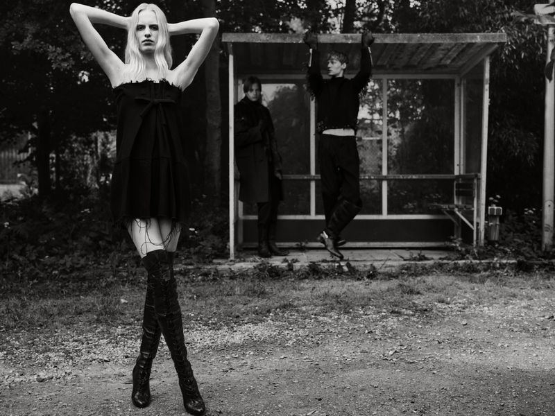 Marrit Krikke in a black strapless dress and thigh-high boots posing outdoors in front of a bus stop shelter with two male models.