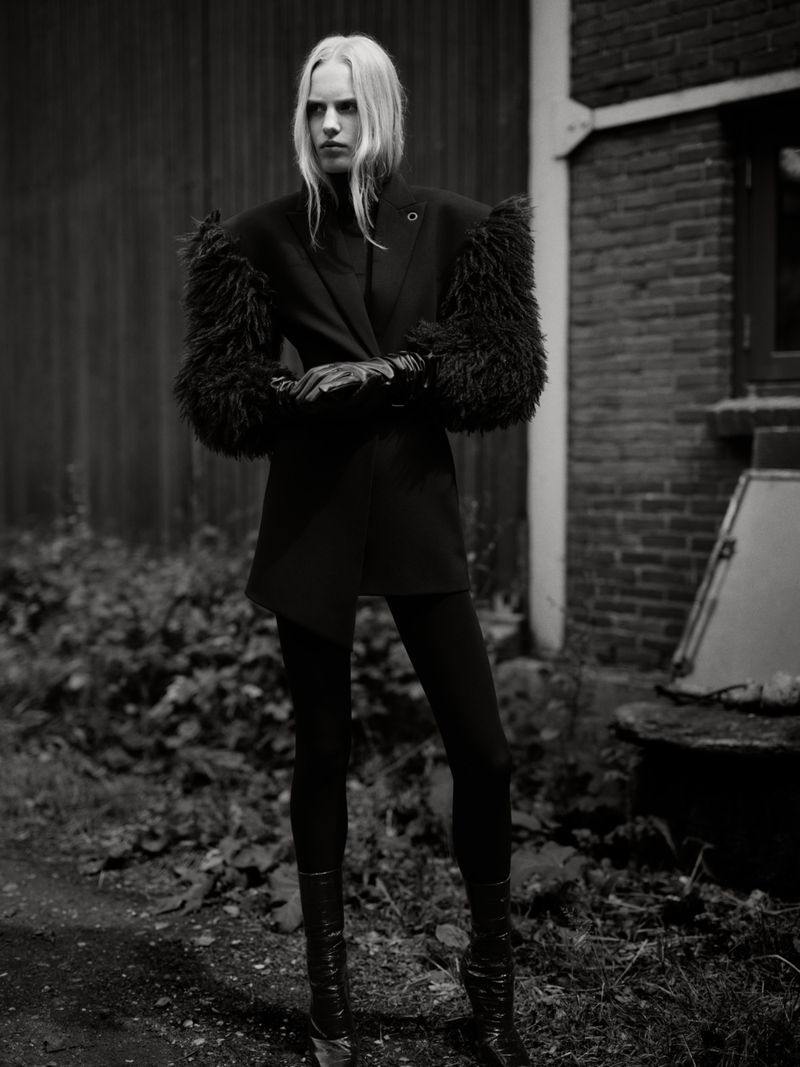 A stylish woman in a black jacket with fur sleeves, black leggings, and boots poses outdoors in a black and white fashion photograph.