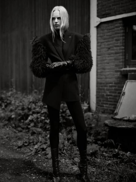 A stylish woman in a black jacket with fur sleeves, black leggings, and boots poses outdoors in a black and white fashion photograph.