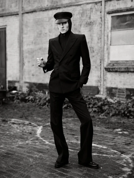 Full-length black and white editorial portrait of a model in a black double-breasted suit and cap standing outdoors before a textured building
