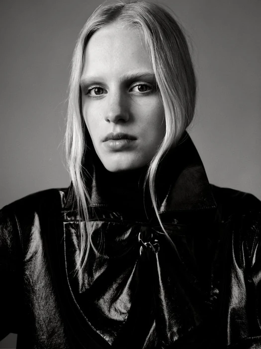 Portrait of a blonde model wearing a dark glossy coat against a neutral grey studio background.