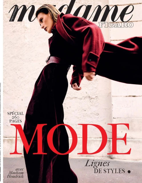 Fashion magazine cover with a woman in a dark red coat and pants standing near a textured wall, featuring large red font.