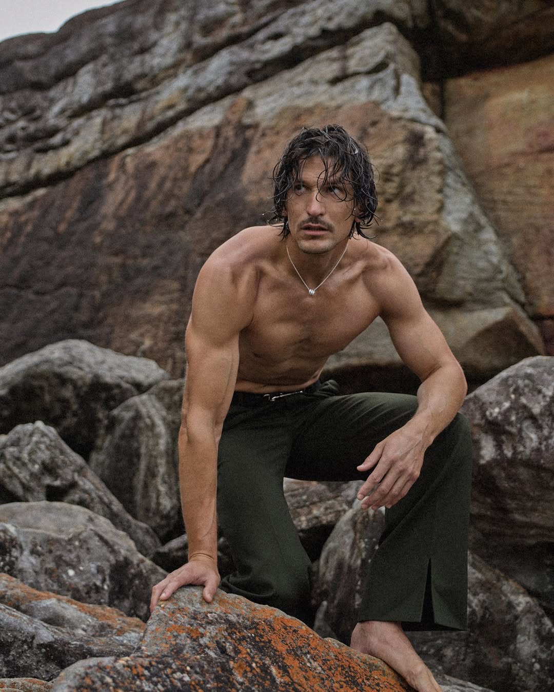 A shirtless man with dark hair and olive pants poses on rocks near the ocean, showcasing a fashion or lifestyle moment in natural light.