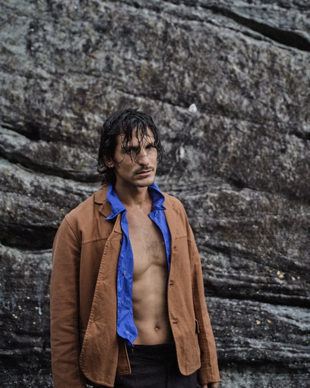 Man in unbuttoned brown jacket stands against a textured rock wall, showing a blue top, dark pants, and a serious facial expression.