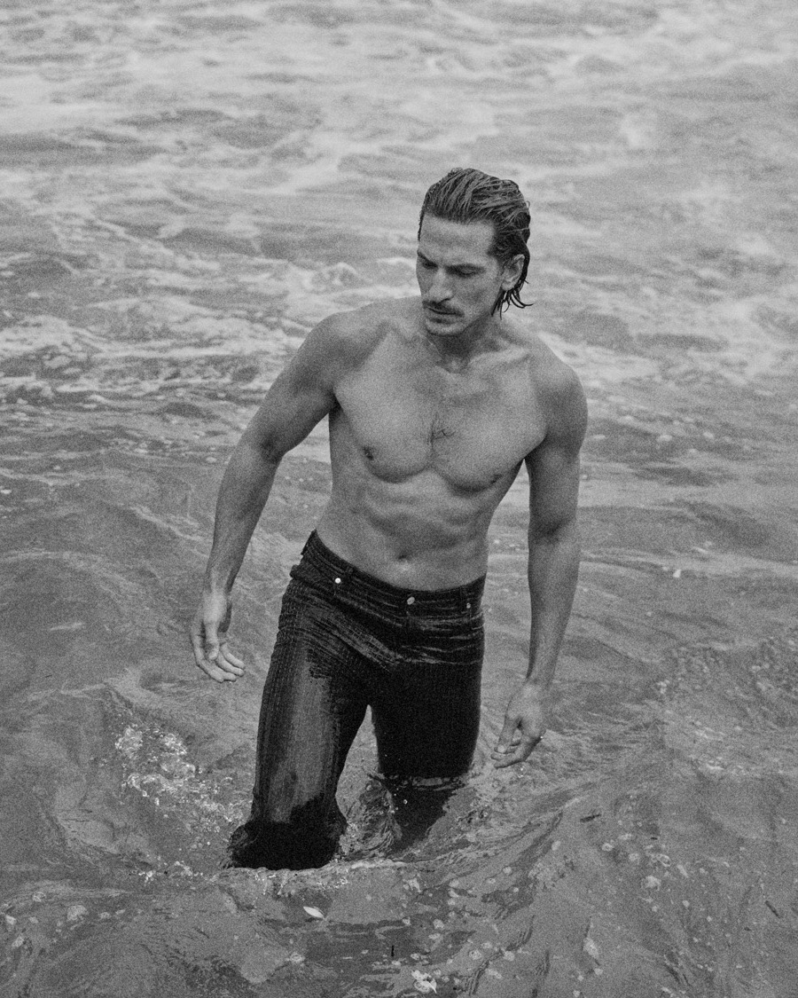 Monochrome portrait of a muscular man in dark jeans standing in ocean waves. Wet hair and water droplets enhance the black and white aesthetic.