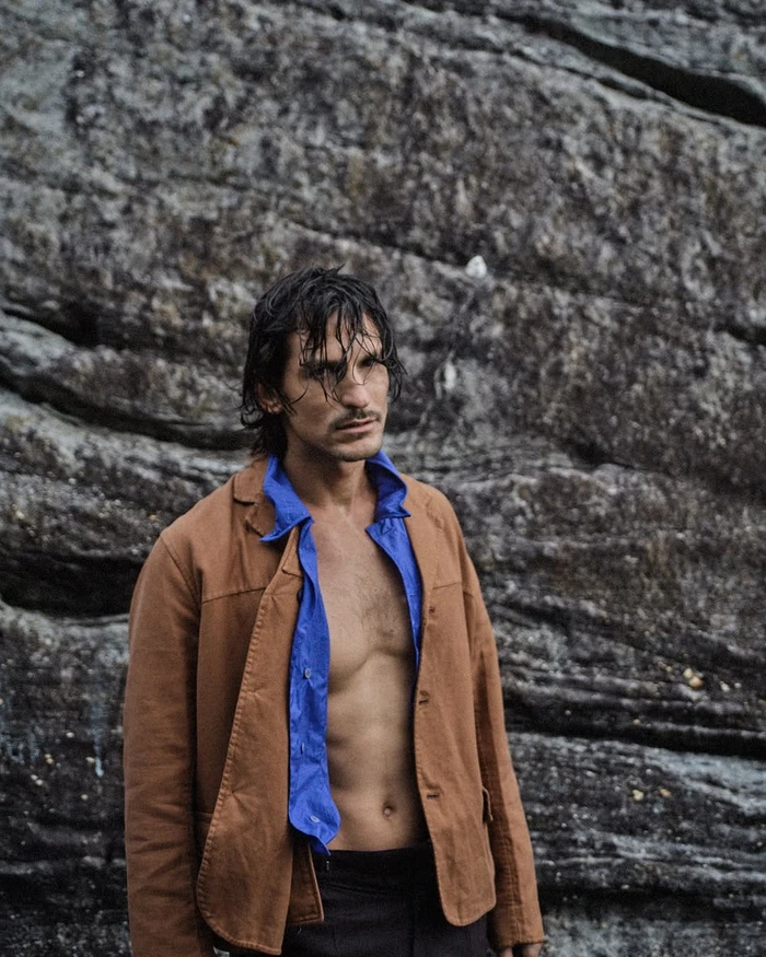 Wet-haired man wearing an open camel jacket and blue shirt against a rocky background