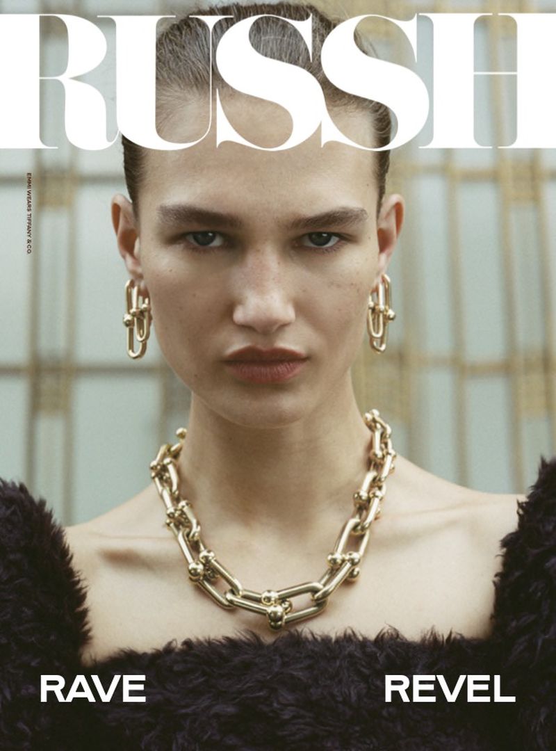 Close-up of a fashion model wearing gold jewelry, including a necklace and earrings, against a blurred background; magazine cover.