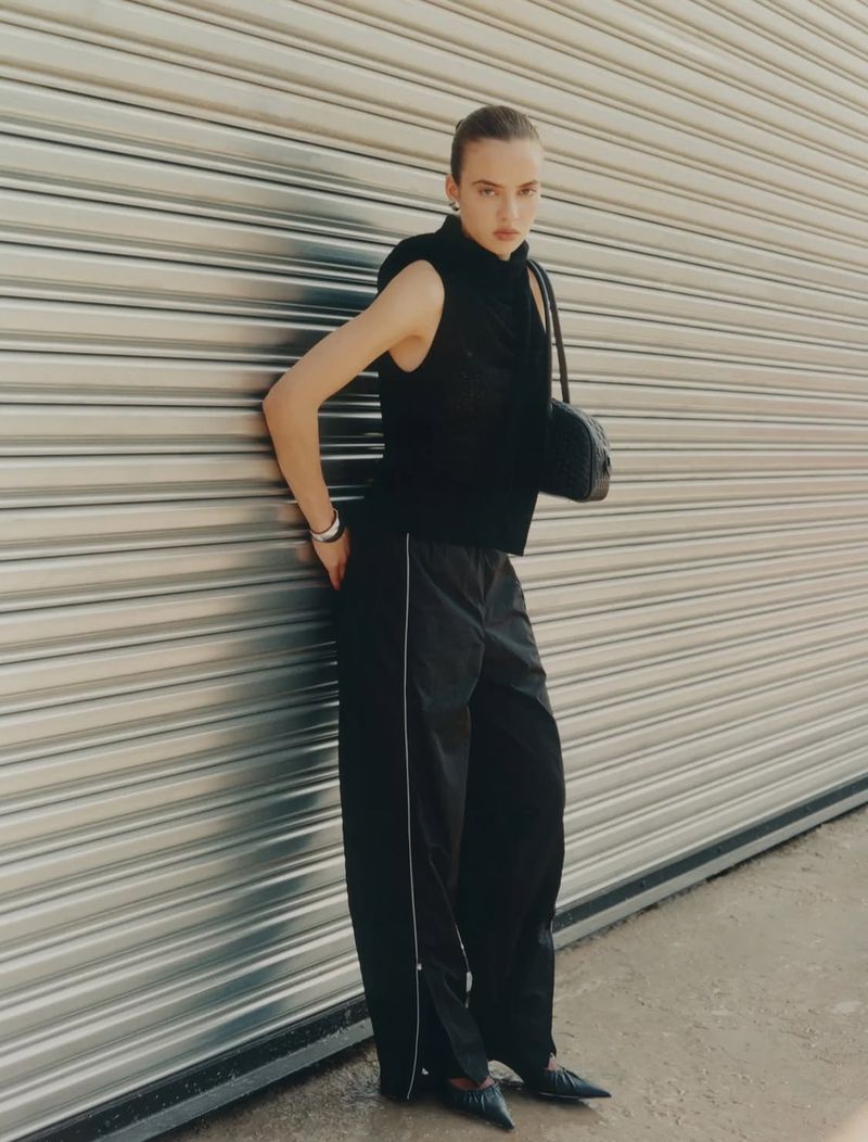 Fashionable young woman with black sleeveless top and track pants leaning against corrugated metal wall with a shoulder bag and pointy shoes.