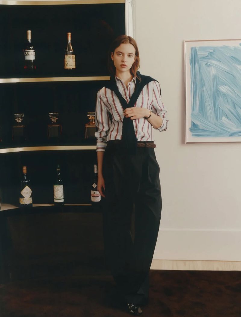 Indoor fashion shot featuring a stylish adult female model in a striped shirt, sweater, and black trousers, with a liquor cabinet backdrop.