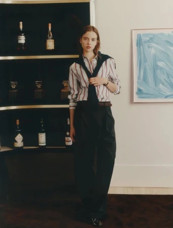 Indoor fashion shot featuring a stylish adult female model in a striped shirt, sweater, and black trousers, with a liquor cabinet backdrop.