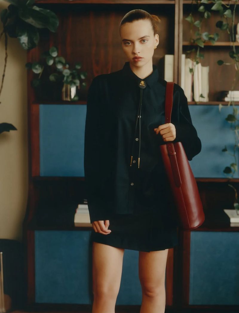 Adult female in a button-down shirt and short skirt carrying a leather tote bag stands indoors, posing in front of a shelf with plants.