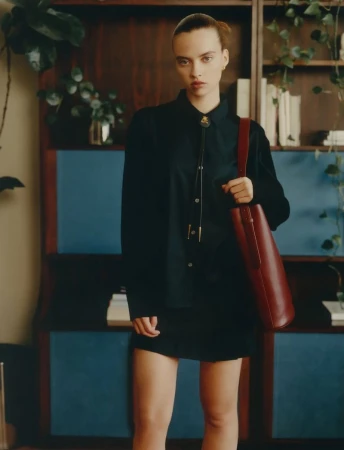 Adult female in a button-down shirt and short skirt carrying a leather tote bag stands indoors, posing in front of a shelf with plants.