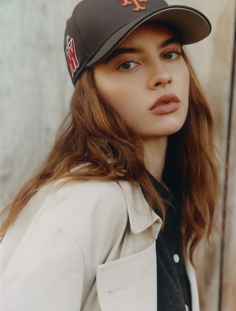 Woman wearing a dark embroidered baseball cap and a beige trench coat looking directly at the camera.