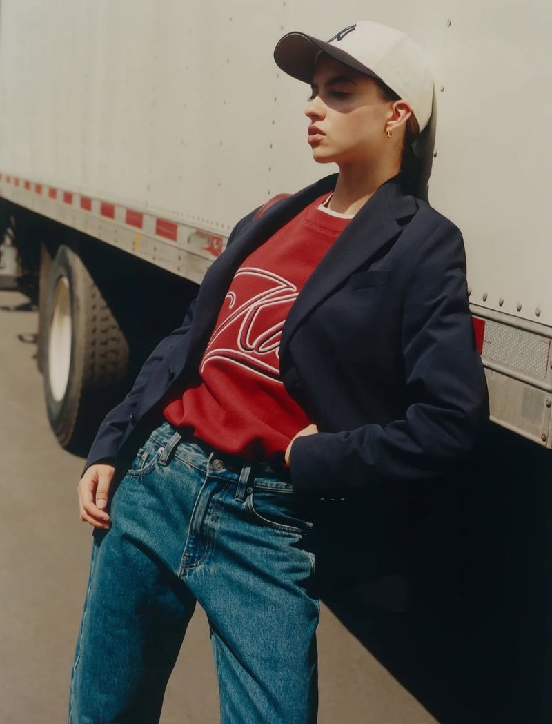 An adult female poses in a blue blazer, red sweatshirt, and blue jeans, leaning against the back of a trailer on an overcast day.