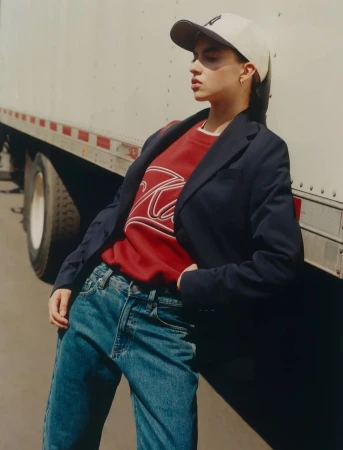 An adult female poses in a blue blazer, red sweatshirt, and blue jeans, leaning against the back of a trailer on an overcast day.