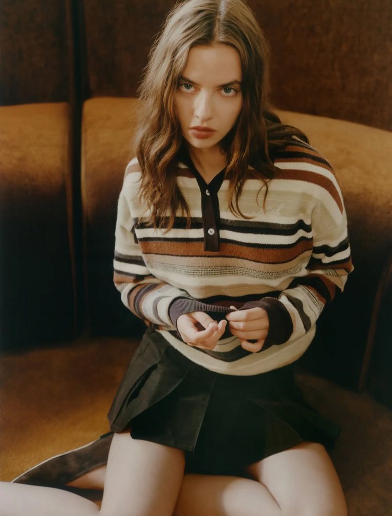 Portrait of a young adult female model with long wavy hair and striped sweater, posing on a brown sofa in an indoor setting.