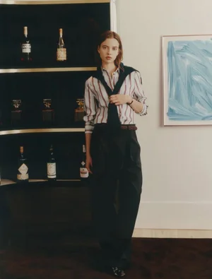 Model wearing a striped button-down shirt, draped black sweater, and dark trousers standing beside a bar shelf.
