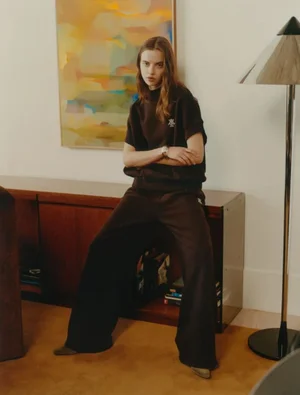 Woman seated on a wooden cabinet wearing a dark brown sweatshirt and matching trousers in a living room interior.