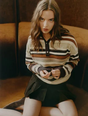 Young woman wearing a multi-toned striped knit polo sweater and dark pleated skirt seated on a brown upholstered booth.