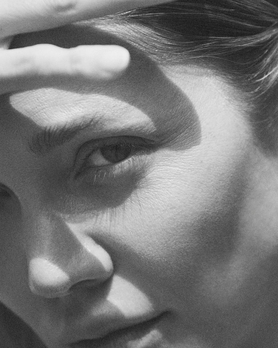 Monochrome close-up portrait of a woman shielding her face with her hand, casting shadows across her eye and nose.