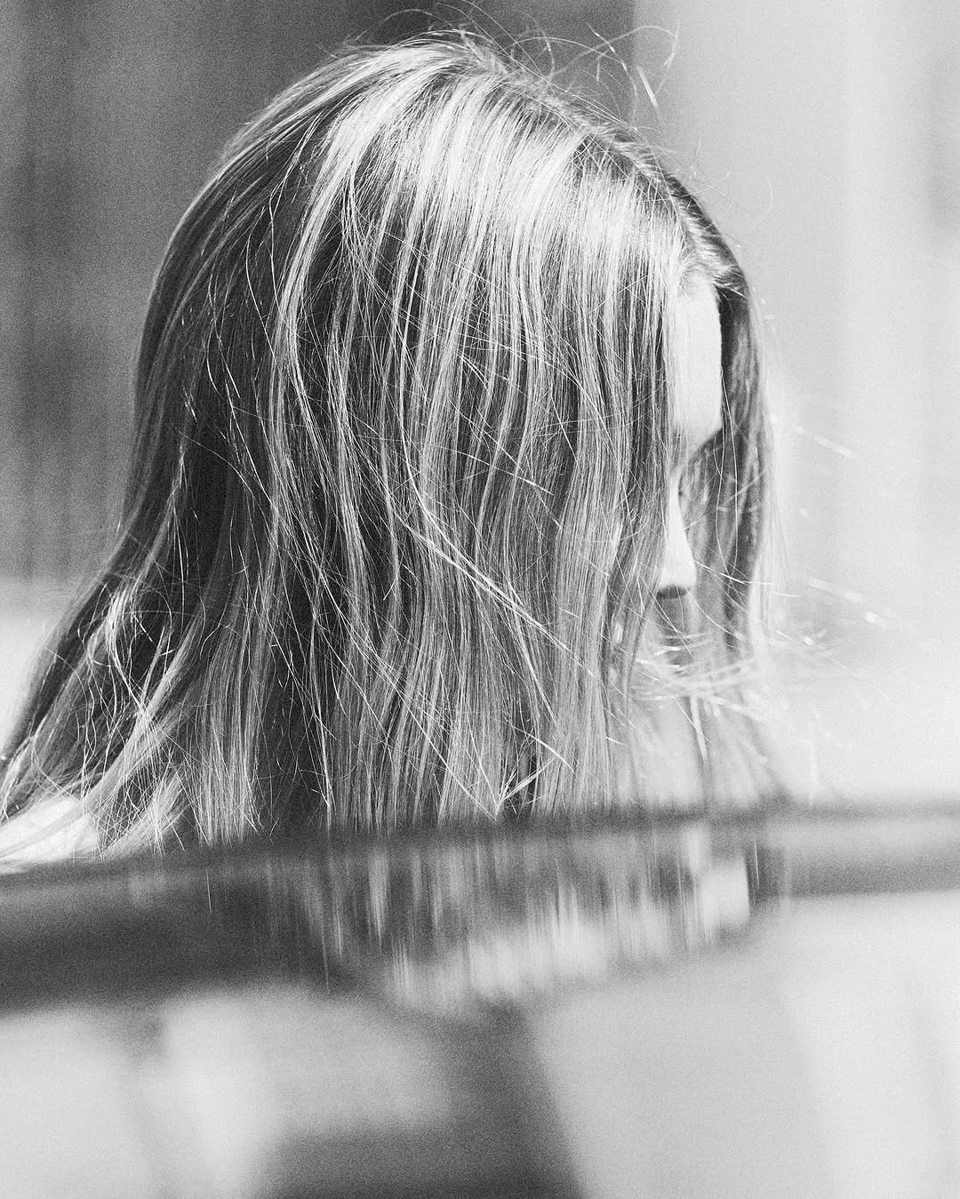 Monochrome portrait of an adult female with long wavy hair partially covering her face, capturing a candid moment in an indoor setting.