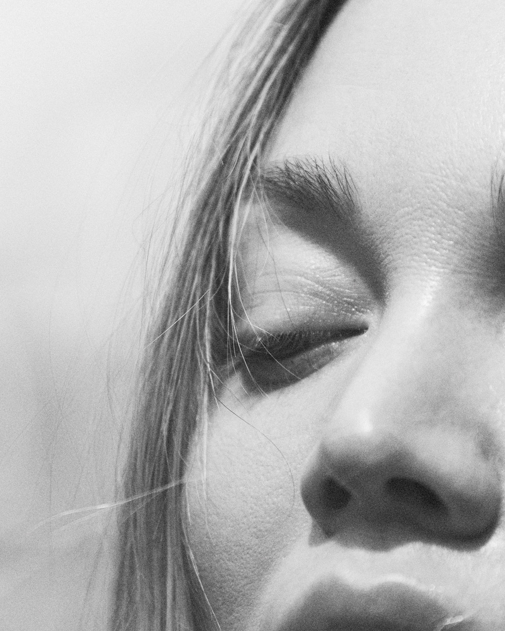 Close-up black and white portrait of a woman with blonde hair and closed eyes, featuring minimalist composition and bright, diffused lighting.