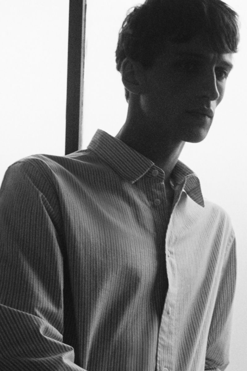 Black and white portrait of a young adult man in a striped dress shirt, standing indoors near a window, with natural lighting.