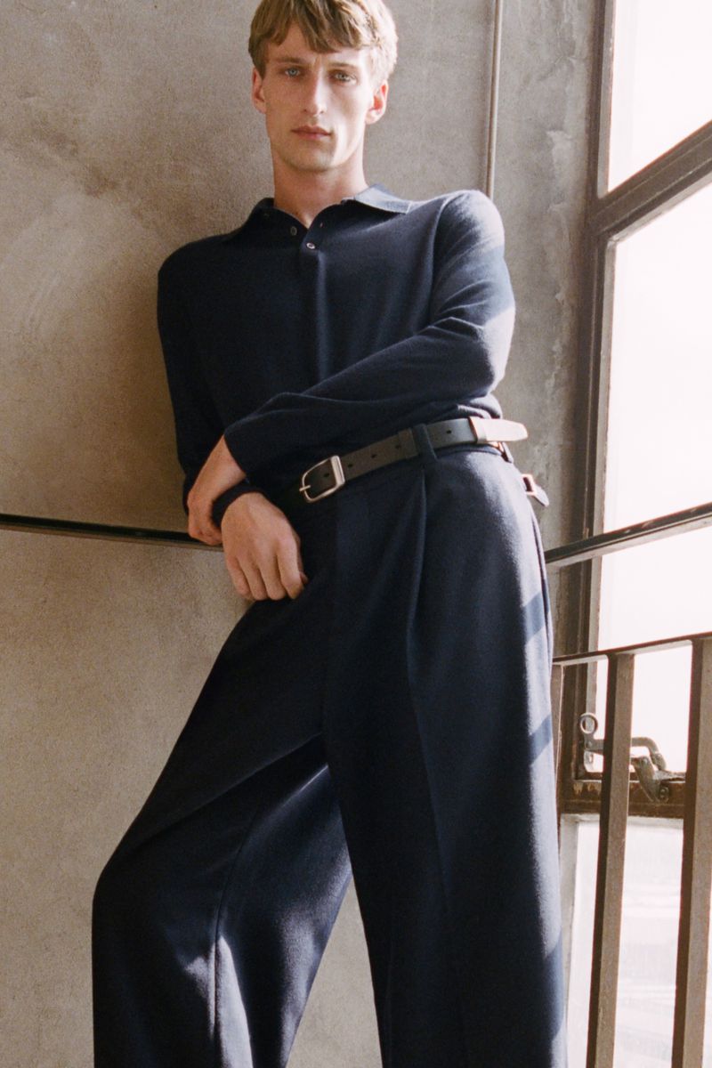 A blond man wearing a navy polo shirt and navy trousers, leaning against a wall with a window with metal bars, indoor setting.